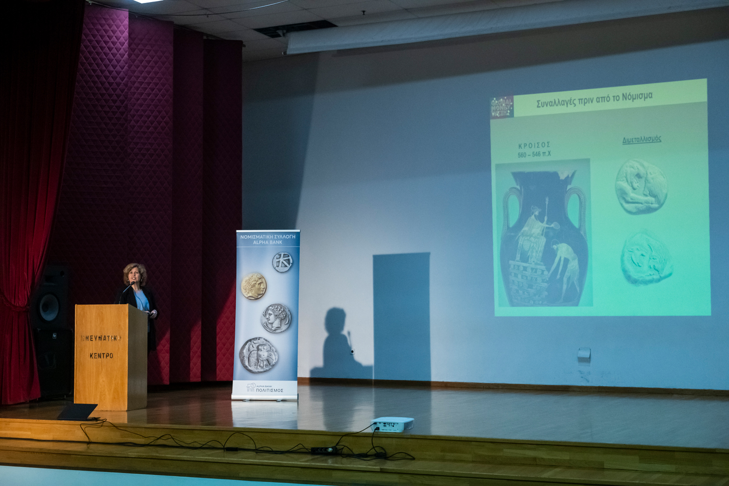 On the left Dr Dimitra Tsangari on the podium and on the right the presentation on a screen. Presentation at the Kyriakopouleio Cultural Centre in Itea