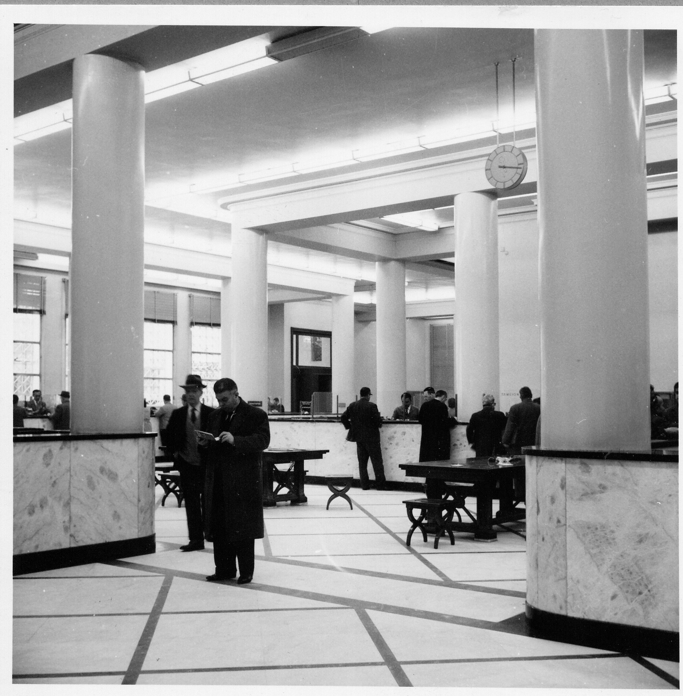 Black-and-white photo of Emporiki Bank’s main hall. It depicts customers and employees.  The main hall of Emporiki Bank’s headquarters