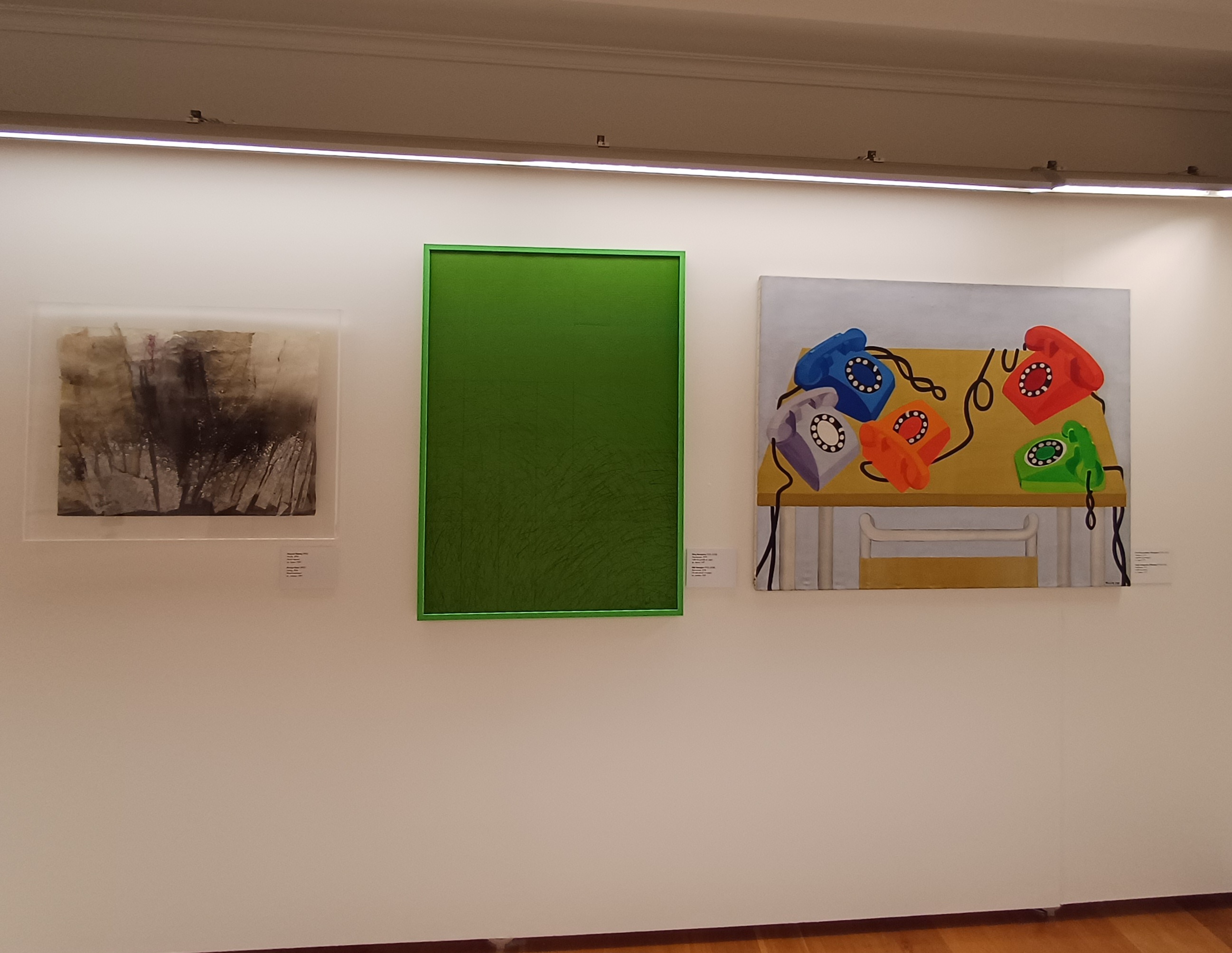Σε λευκό τοίχο, αριστερά, αφηρημένος πίνακας, στη μέση πράσινος πίνακας και δεξιά πίνακας με πολύχρωμα τηλέφωνα. Φωτογραφία από τους χώρους της έκθεσης