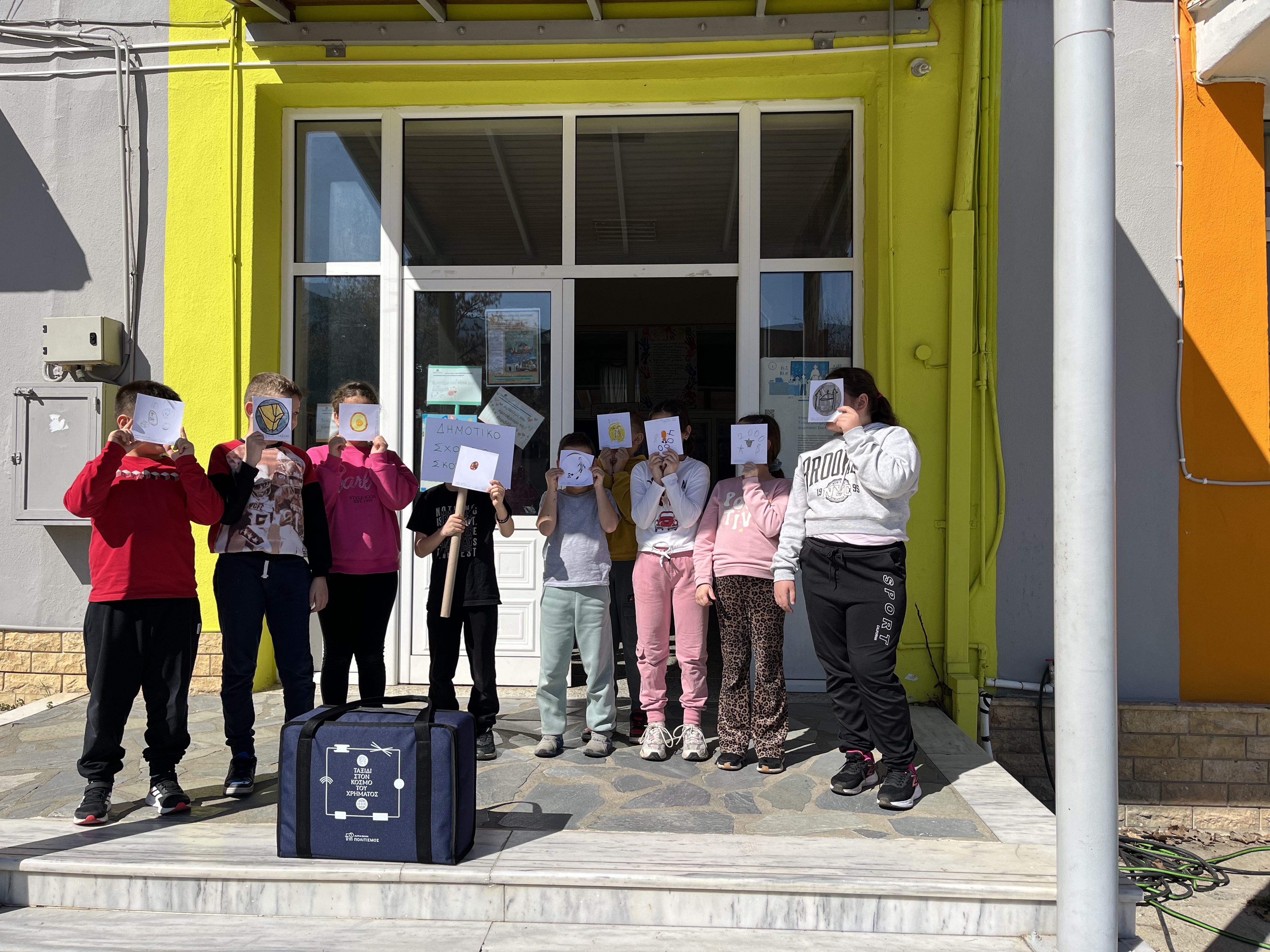 Children at a school entrance holding their creations in front of their faces. At the front, the museum kit. Snapshot from educational programme