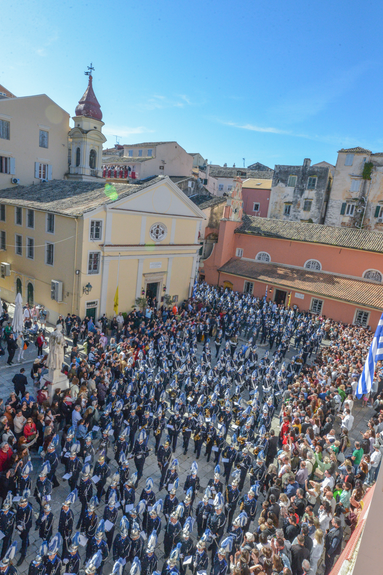 Musicians of the Mantzaros Philharmonic Society in blue uniforms and hats marching through the crowded square. The Mantzaros Philharmonic Society