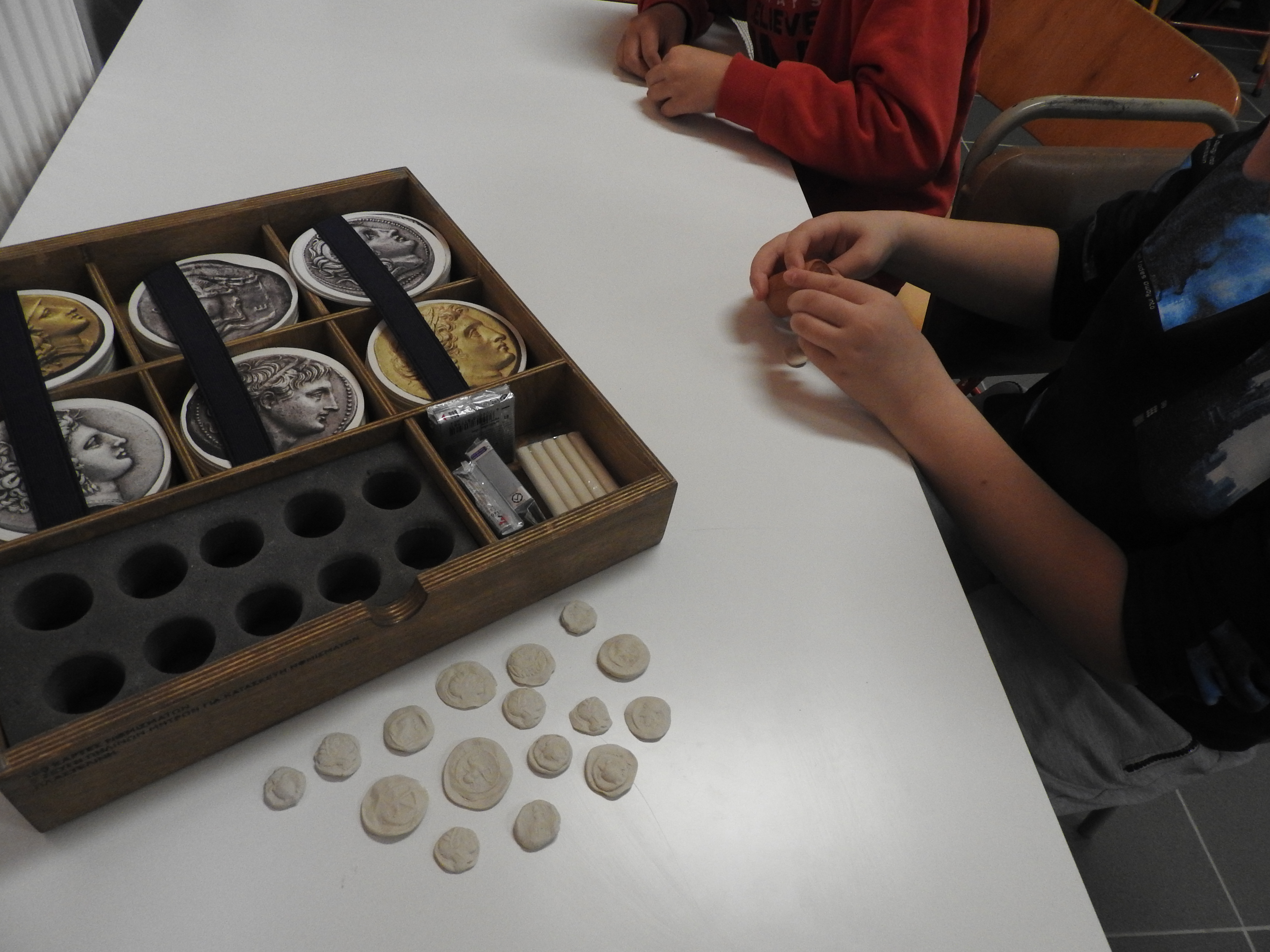 On a desk, a wooden case with coin replicas. In front of it, 2 children make coins out of play dough. Snapshot from educational programme