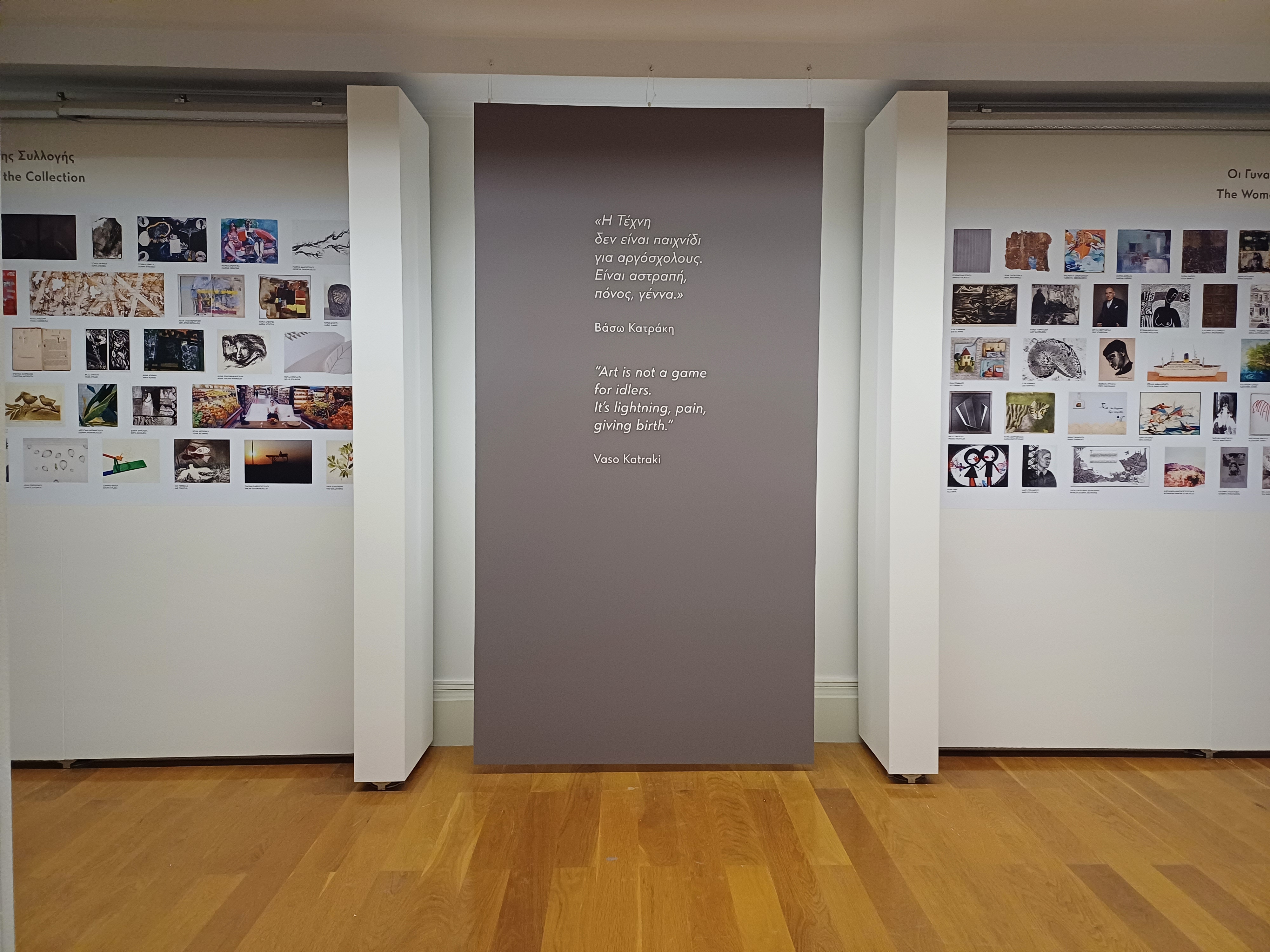 Πολύχρωμες φωτογραφίες σε λευκό τοίχο. Στη μέση, μεγάλο καφέ banner με φράση της ζωγράφου Βάσω Κατράκη. Φωτογραφία από τους χώρους της έκθεσης