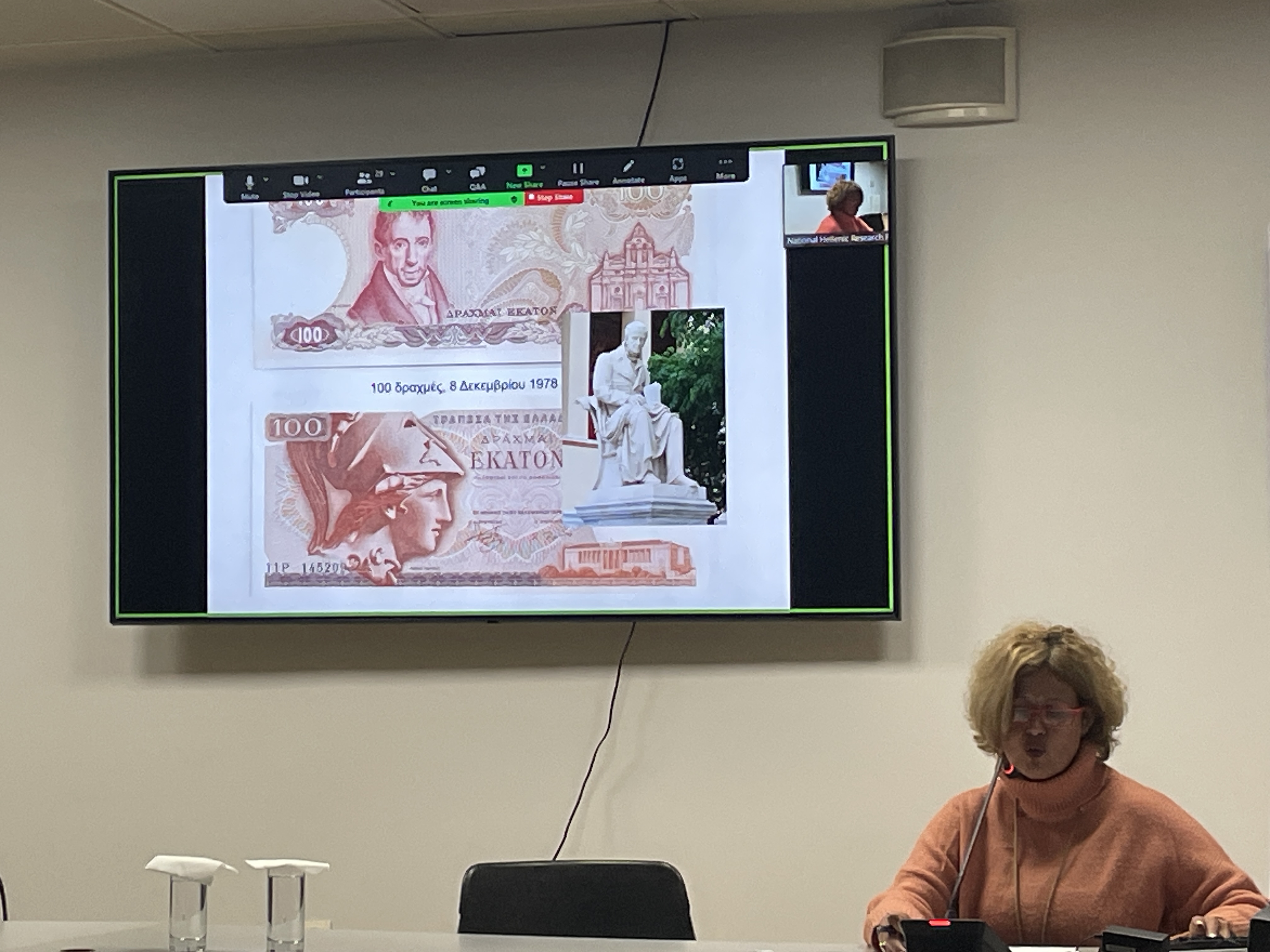 Dr Tsangari speaking seated behind a table. Behind her, a screen displays 100-drachma banknotes. Highlight from the lecture