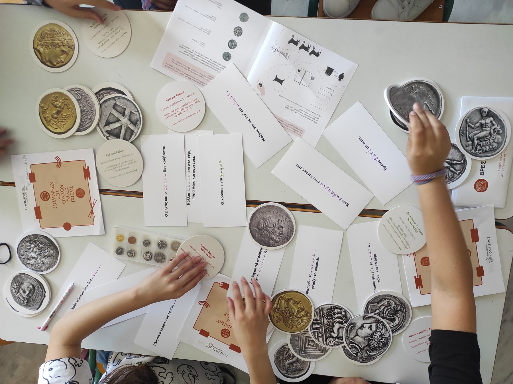 Coin cards, case with coin replicas, and brochures on 2 white desks. Around them, children’s hands. Snapshot from educational programme