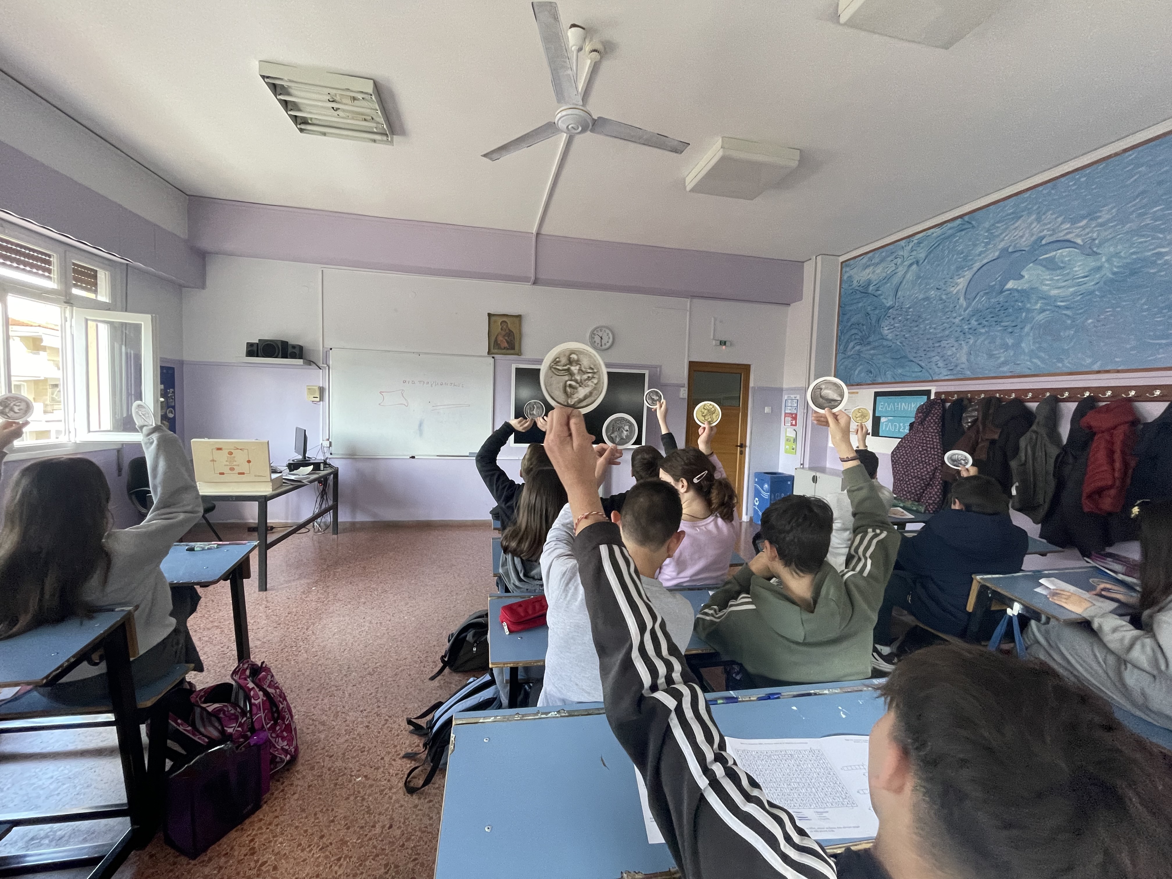 Classroom. Children seated at their desks with their backs to the camera, holding up coin cards. Snapshot from educational programme