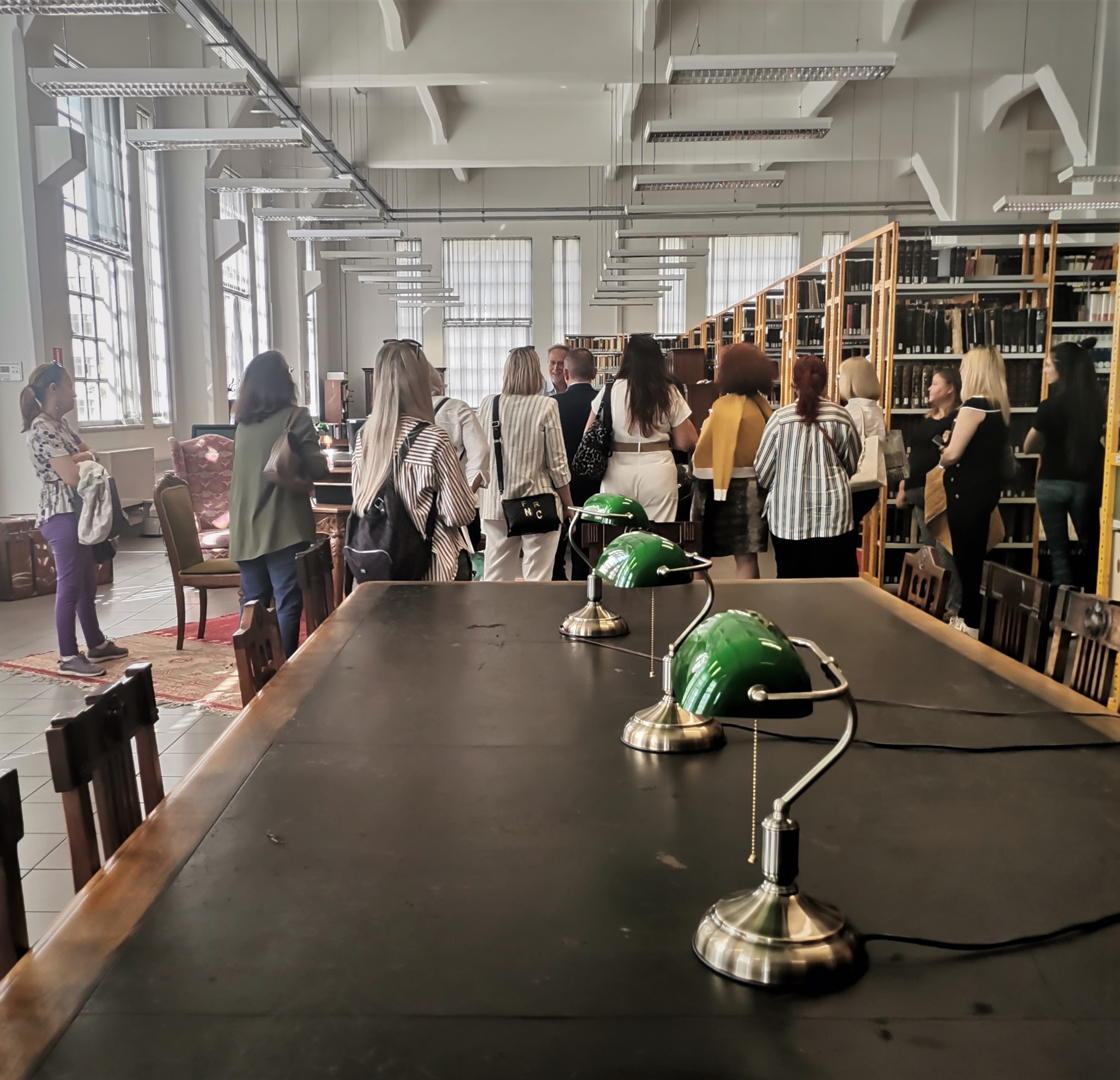 Large black table with green lamps. In front of it, visitors attend the tour next to bookcases.  Highlight from the guided tour