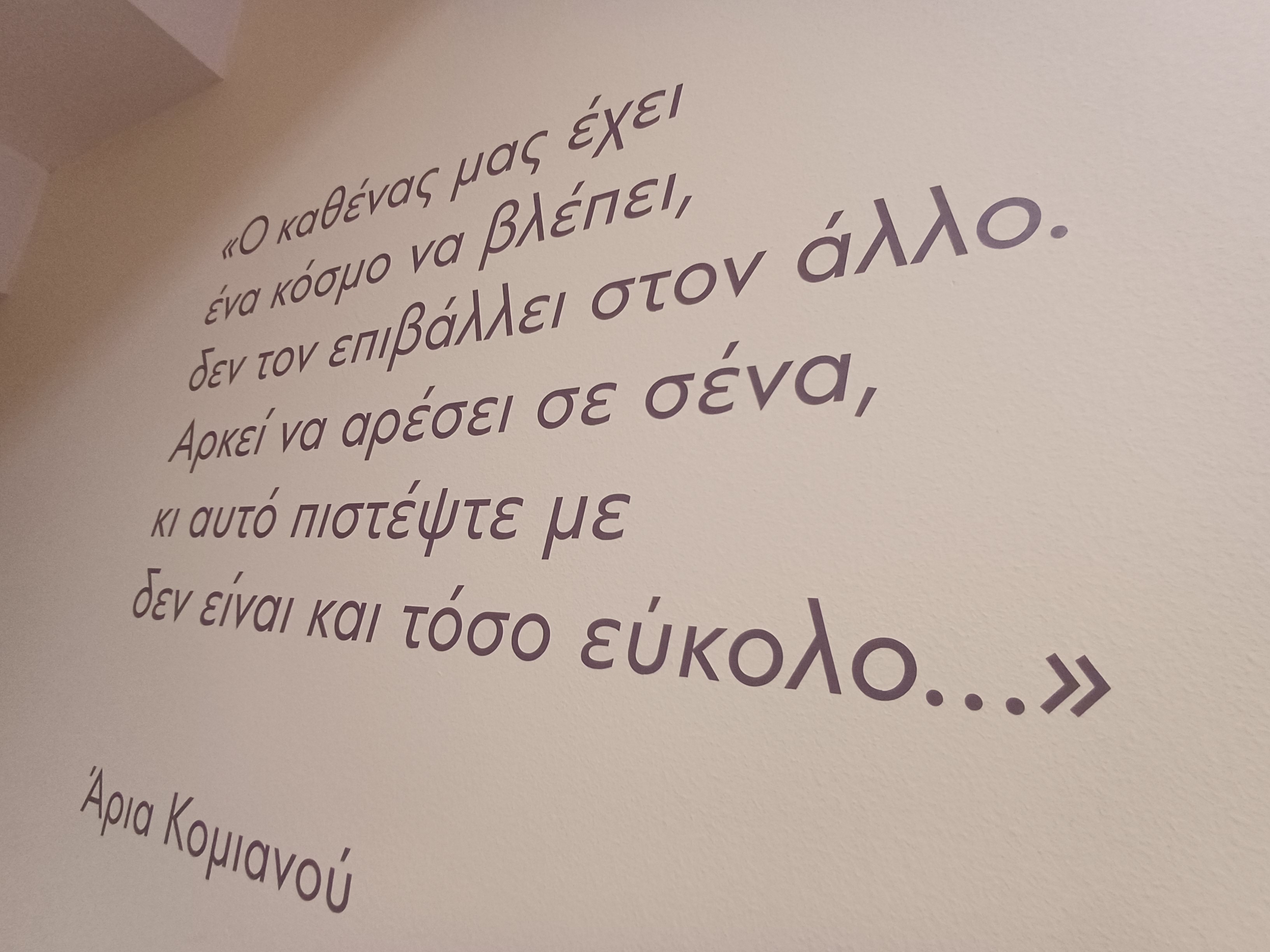 Λευκός τοίχος με εκτύπωση μίας φράσης της χαράκτριας Άριας Κομνηνού με γκρι γράμματα. Φωτογραφία από τους χώρους της έκθεσης