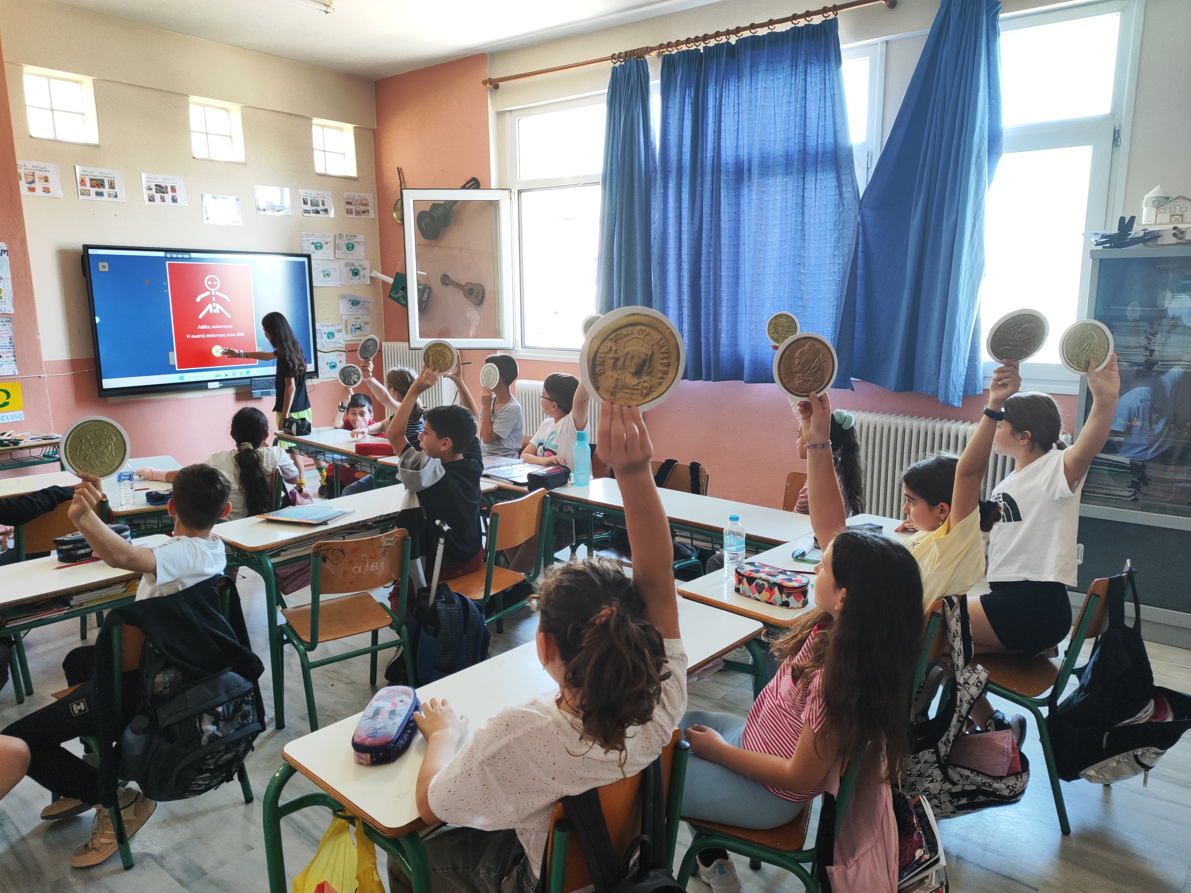 Children holding paper coin replicas and a girl using the interactive whiteboard. Snapshot from educational programme
