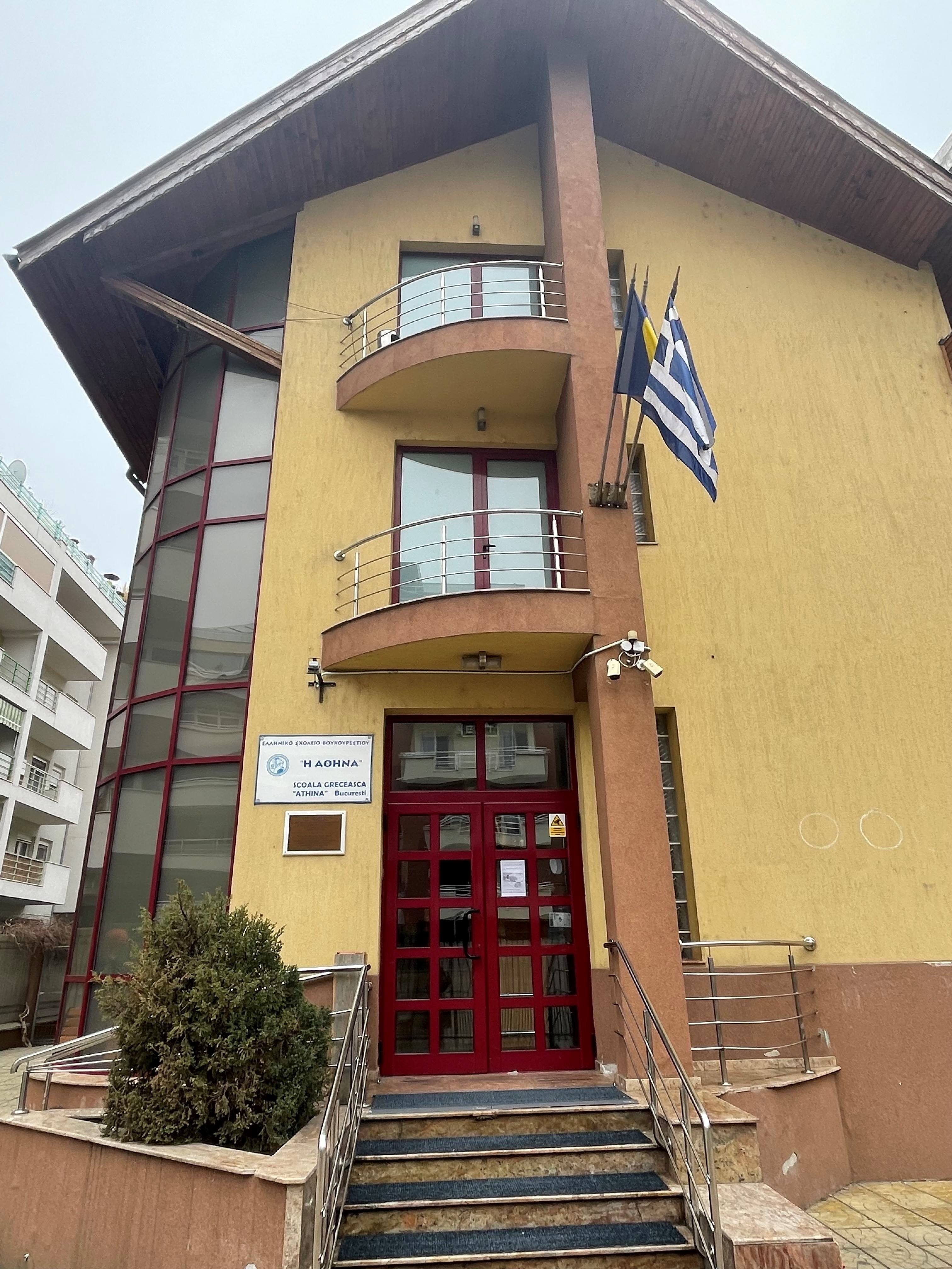 Façade of a two-storey building with a red door, beige walls and the Greek flag on the first-floor balcony. The Greek School of Bucharest
