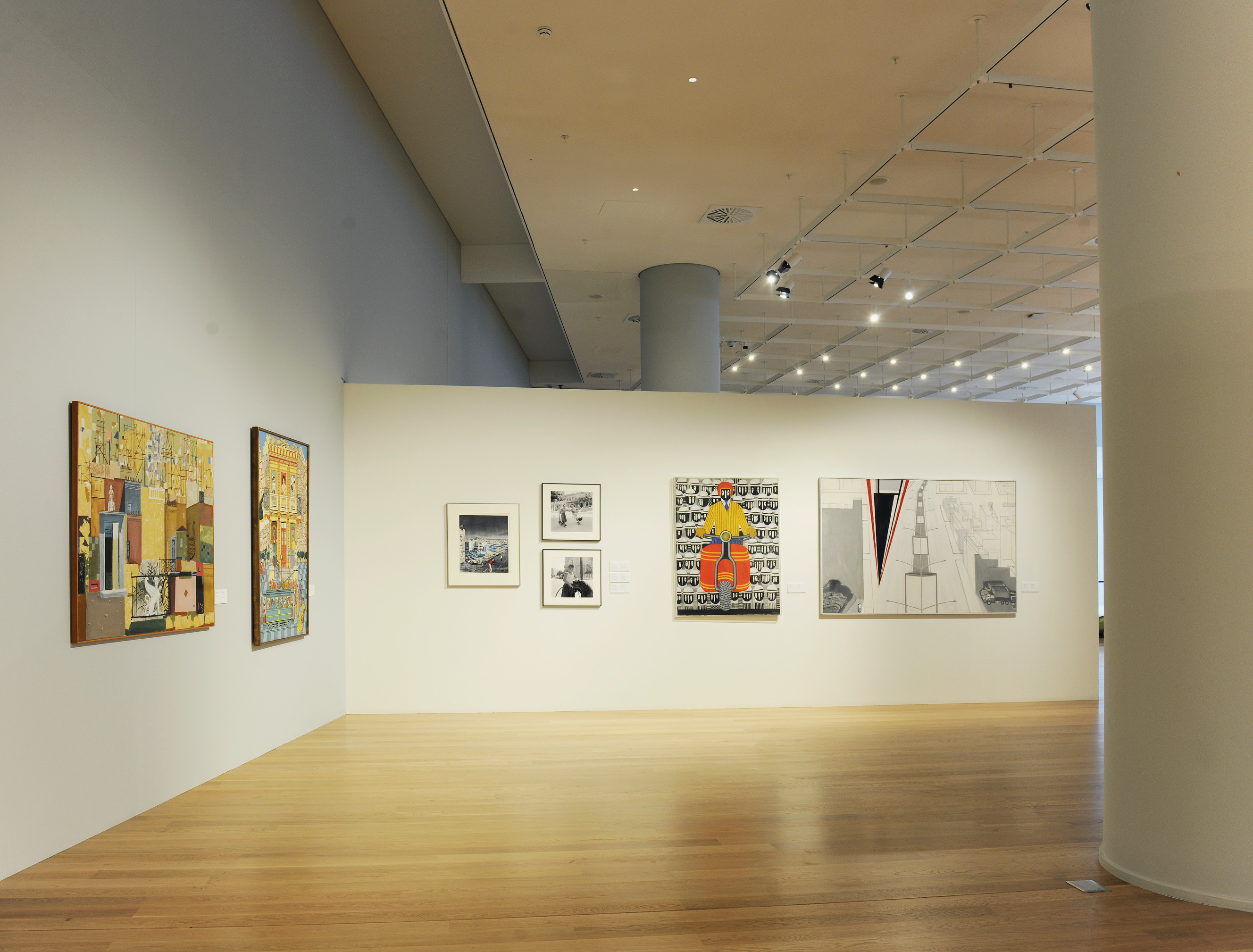 Black and white photos on the left and a painting with a motorbike rider and one with an avenue and blocks of flats on the right. Exhibition area