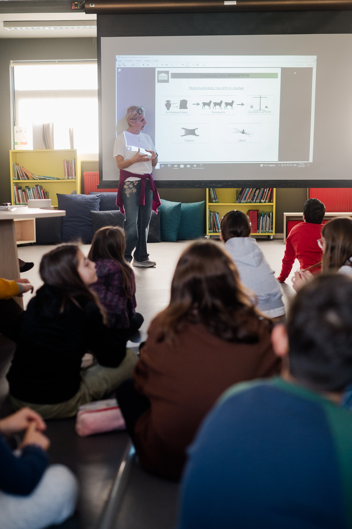 Children sitting on the floor watching a woman speak next to the projector. Highlight from the presentation