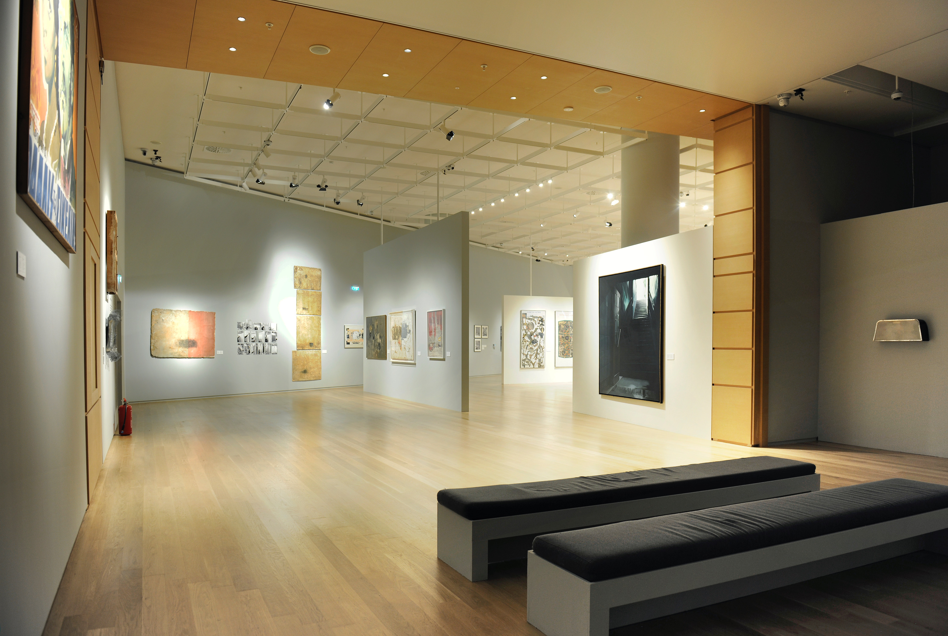 Paintings on the far back wall and a half wall with paintings on both sides, along with a bench with cushions.  Exhibition area