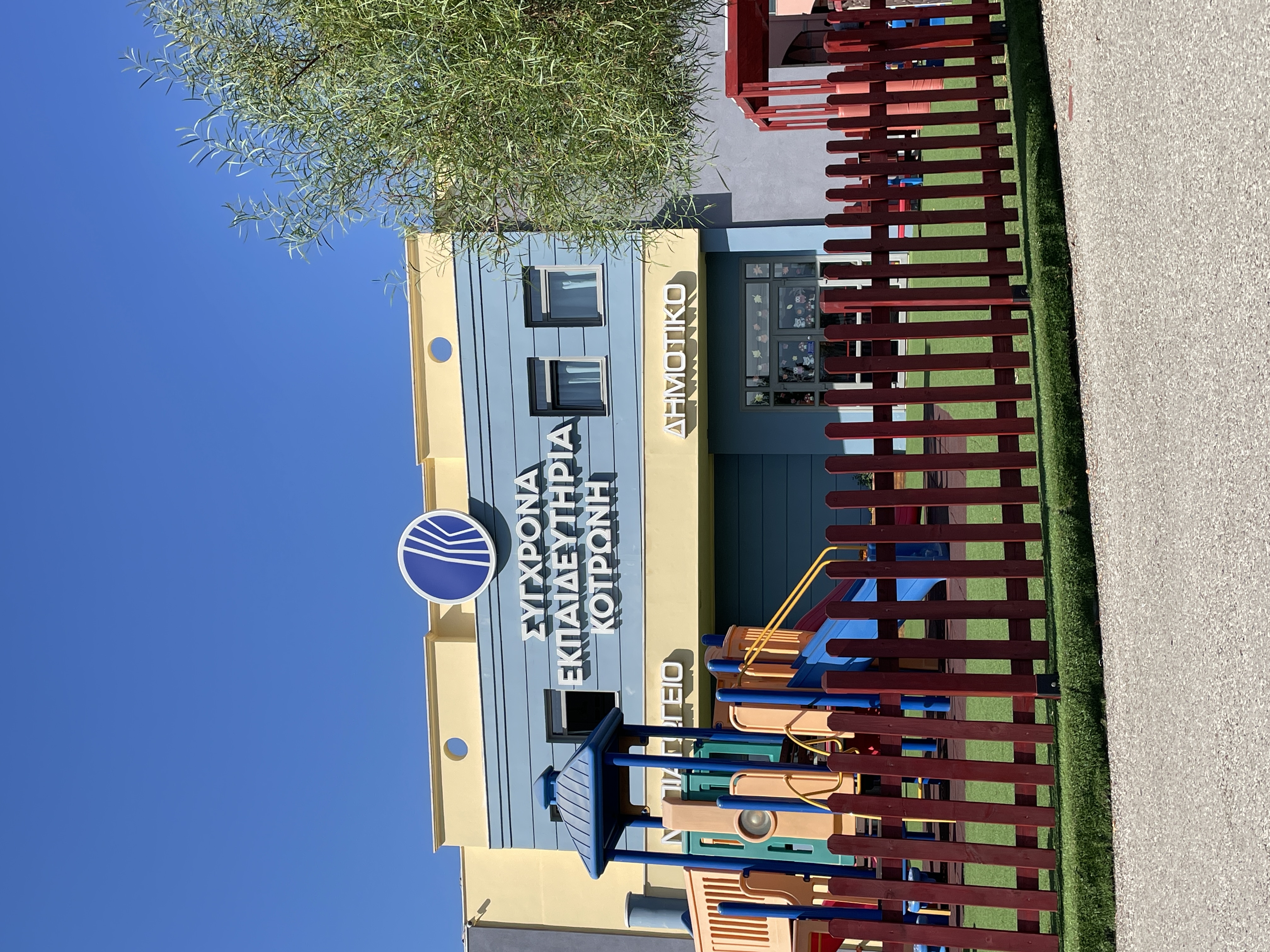 School yard with lawn and a slide and behind a building with the inscription “Modern Kotronis Schools, Kindergarten, Primary School”. Façade of the Kotronis Schools in Nafpaktos