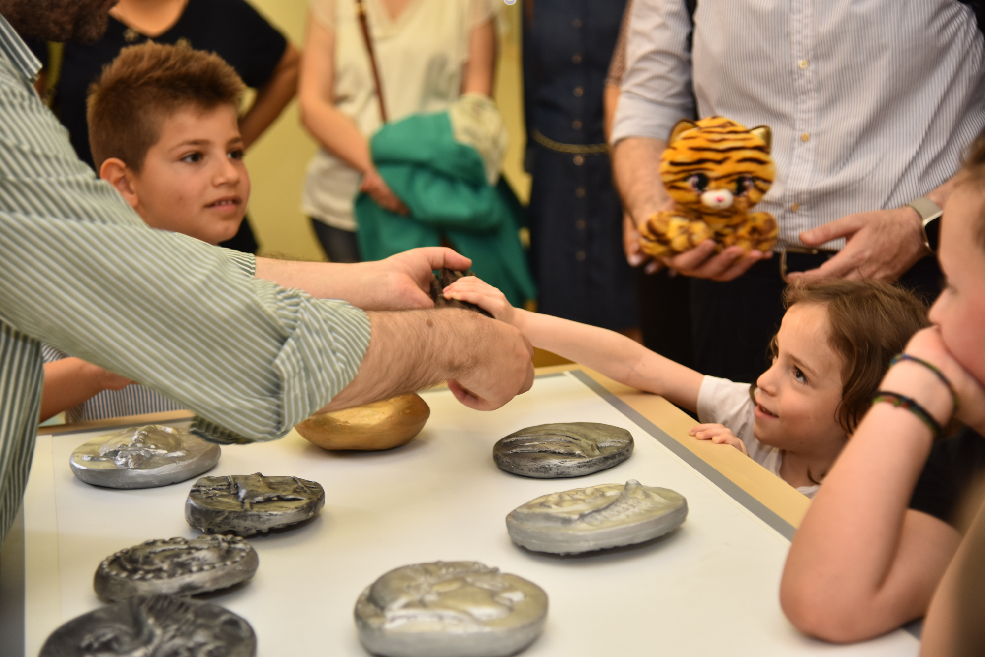 Children and their parents attend a tour around a table with enlarged coin replicas. Snapshot from a touch tour at the Banknote Museum