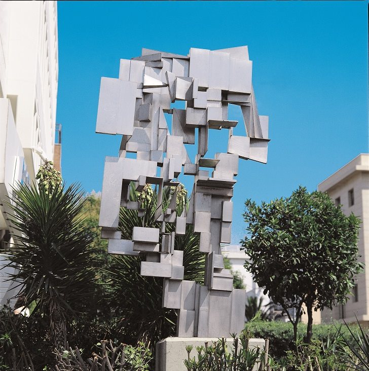 Abstract steel sculpture, resemblant of a tree among plants. Buildings to the left and right. 
