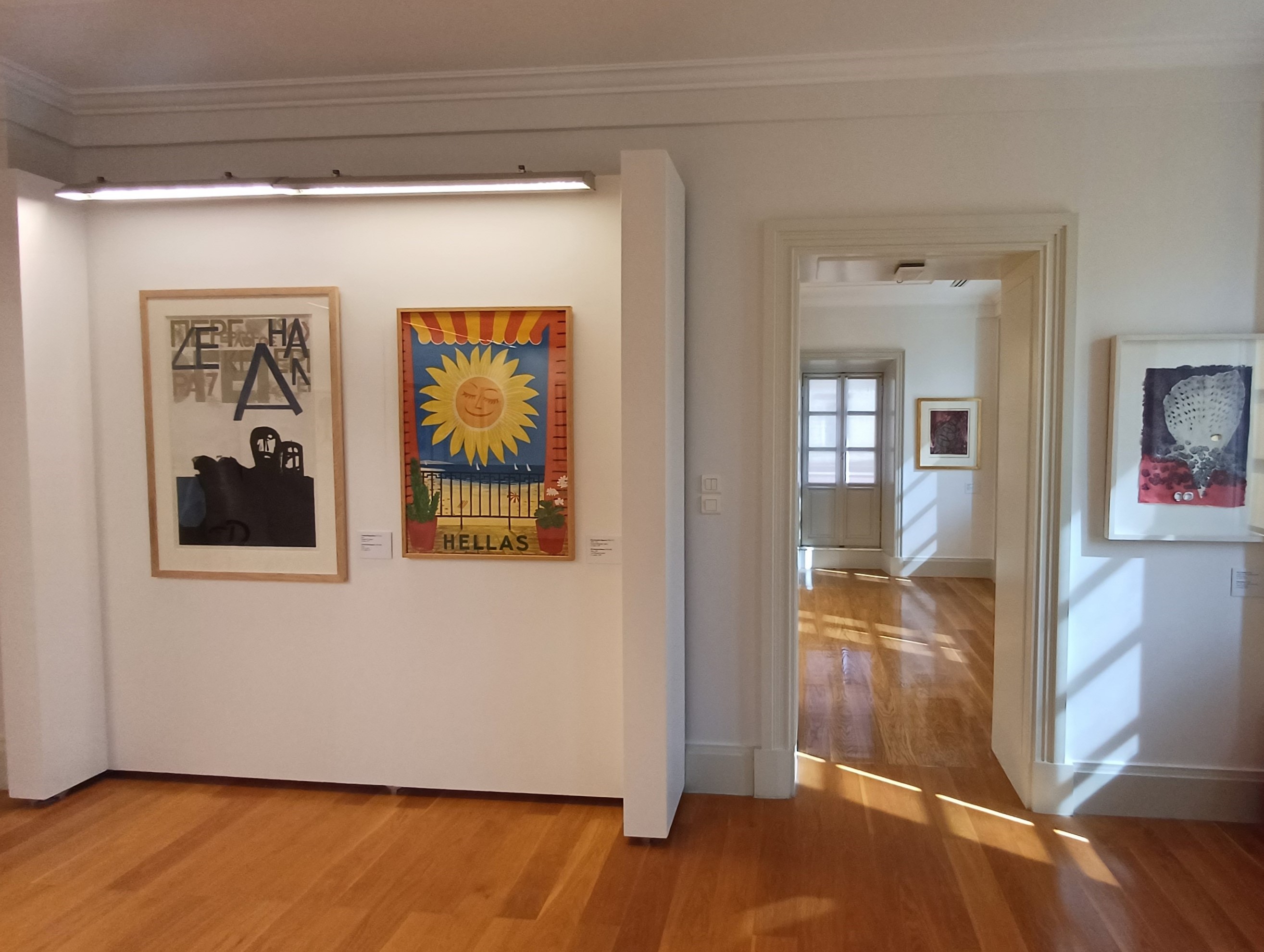 Σε λευκό τοίχο, αριστερά, πίνακας με γράμματα και ανθρώπινες μορφές, δεξιά ένας με ηλιοτρόπιο. Δίπλα, πόρτα. Φωτογραφία από τους χώρους της έκθεσης