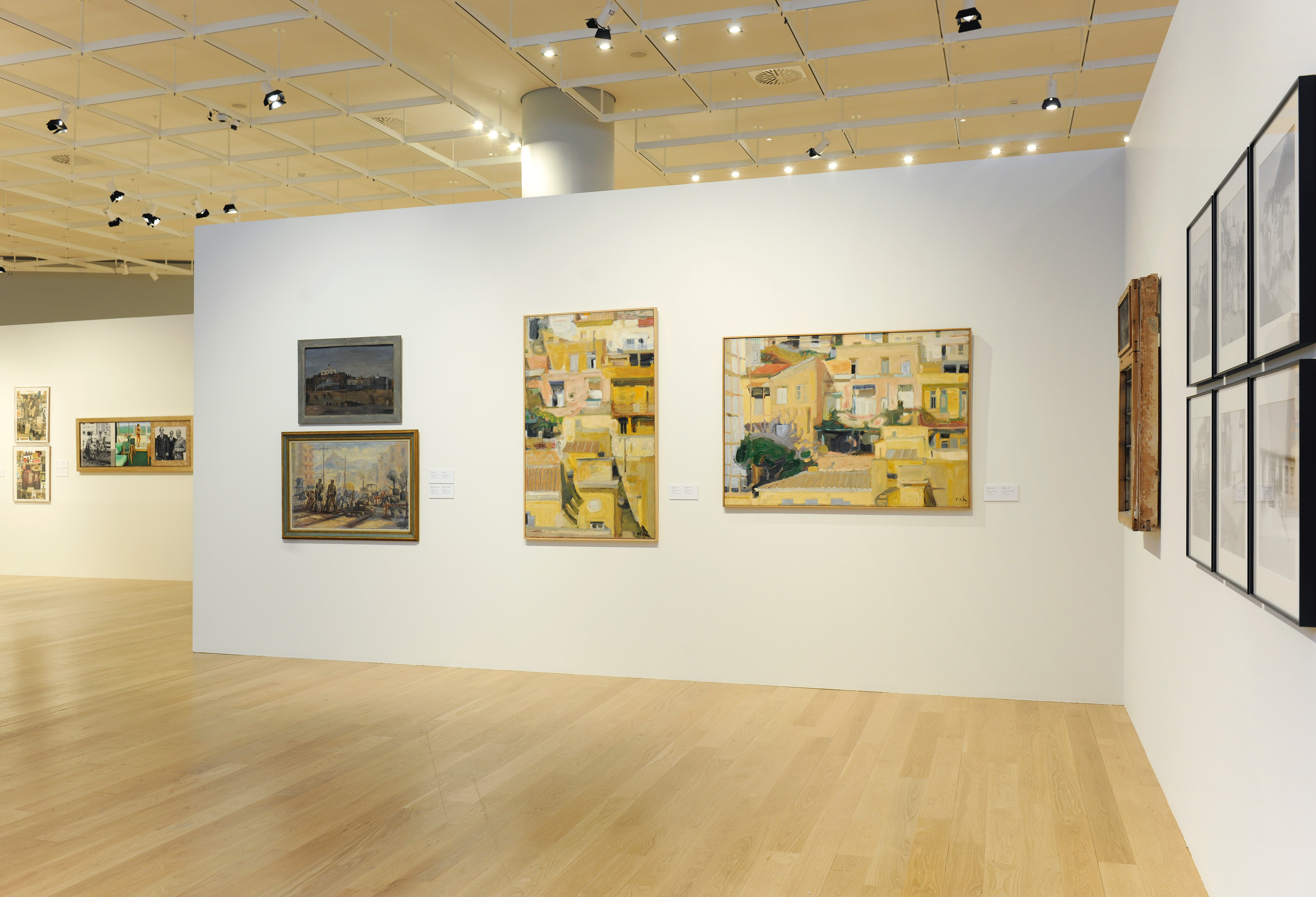 Paintings on a white wall. Painting with city roadworks on the left and multi-coloured paintings with houses on the right. Exhibition area
