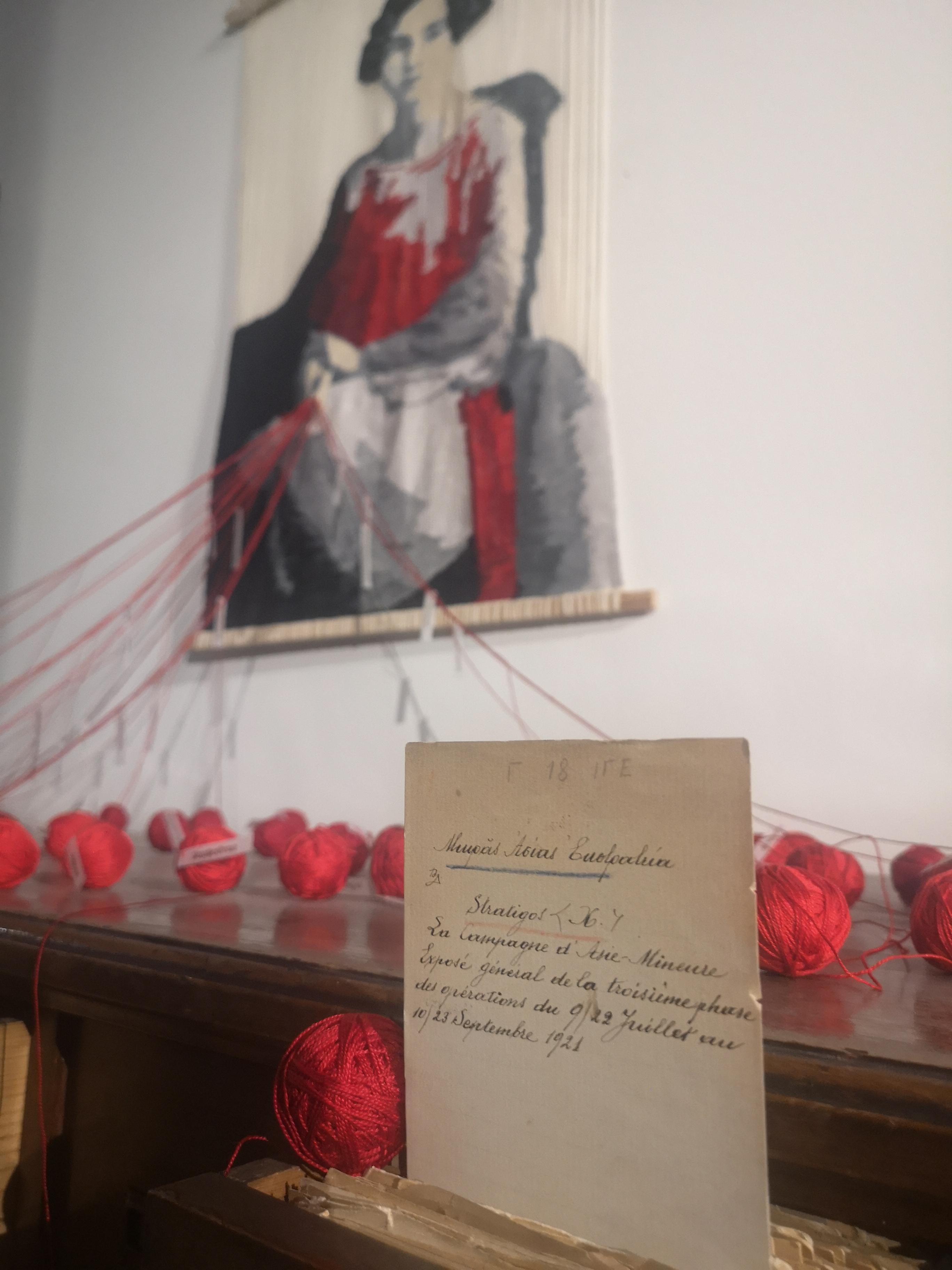 On a white wall, grey and red yarns form a female figure. At the front, balls of red yarns and a note. Exhibit