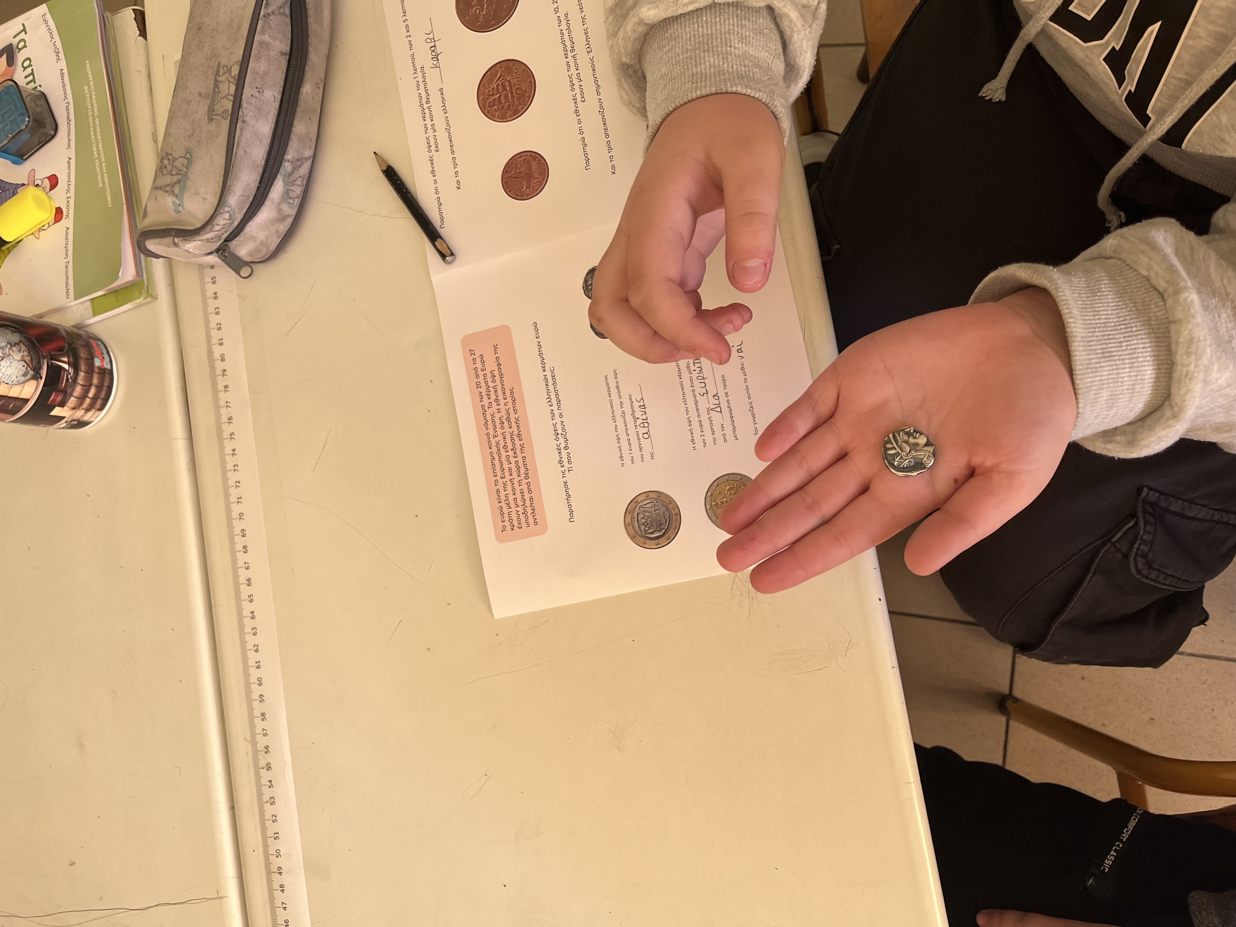 Child holding a coin replica on their palm. On the desk in front of them, a programme brochure. Snapshot from educational programme