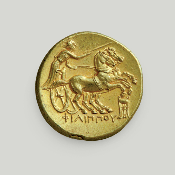 Οπίσθια όψη χρυσού στατήρα Φιλίππου Β΄ με ηνίοχο πάνω σε άρμα με 2 άλογα, τρίποδο και την επιγραφή ΦΙΛΙΠΠΟΥ στο κάτω μέρος.  Οπίσθια όψη χρυσού στατήρα Φιλίππου Β΄, Μακεδονία