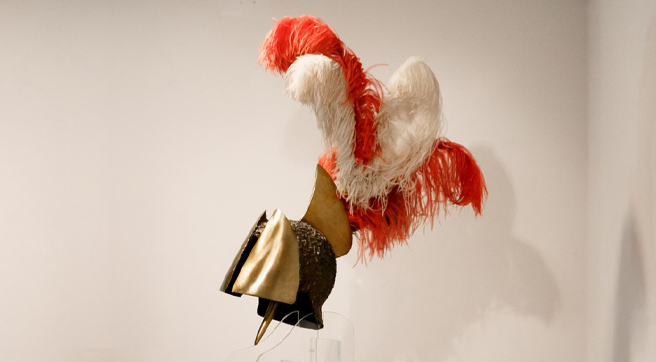 Bronze helmet with feathers, before a white wall, on top of a clear base. Bronze helmet with feathers, before a white wall, on top of a clear base.