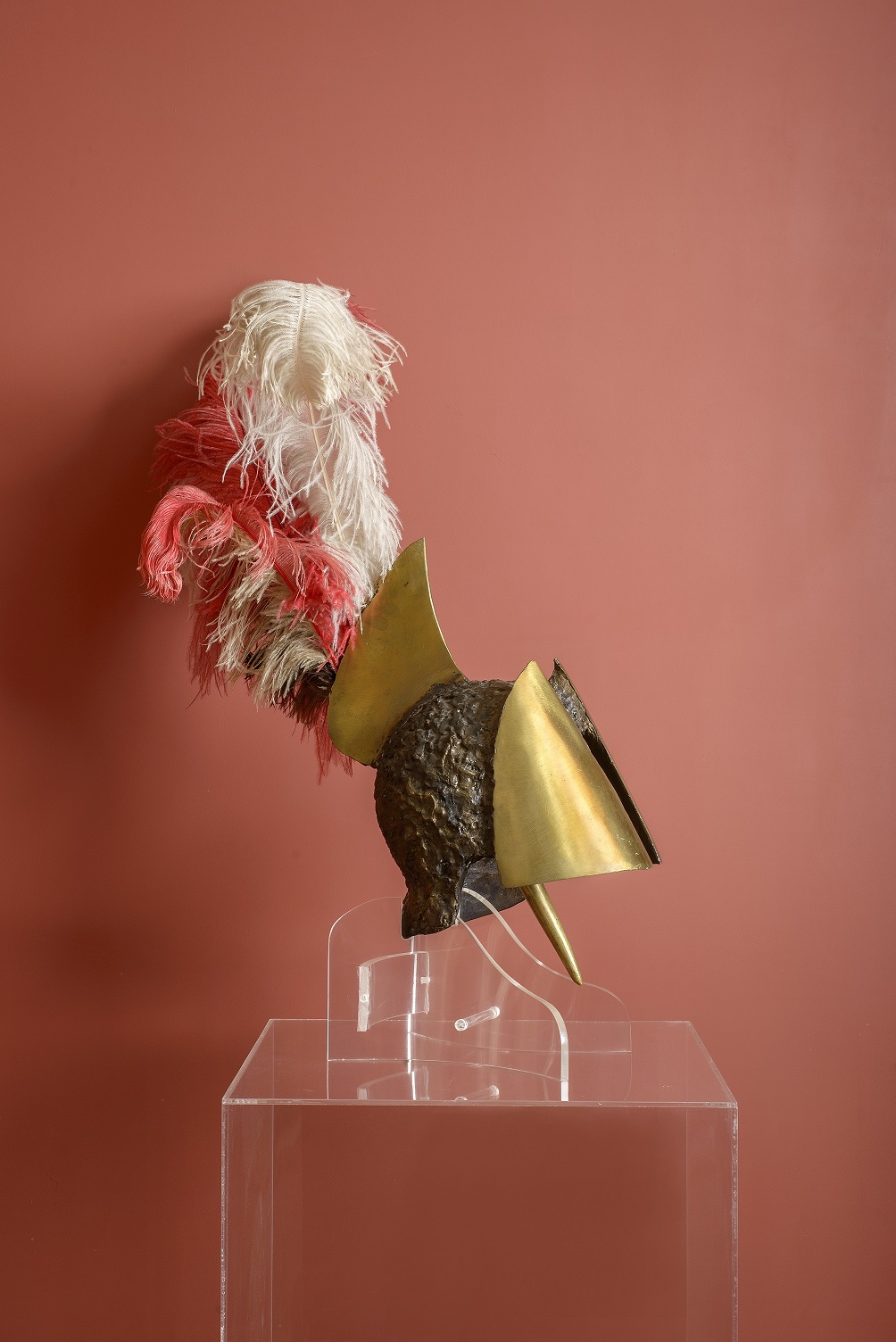 Bronze helmet with feathers, before a red wall, on top of a clear base. Bronze helmet with feathers, before a red wall, on top of a clear base.